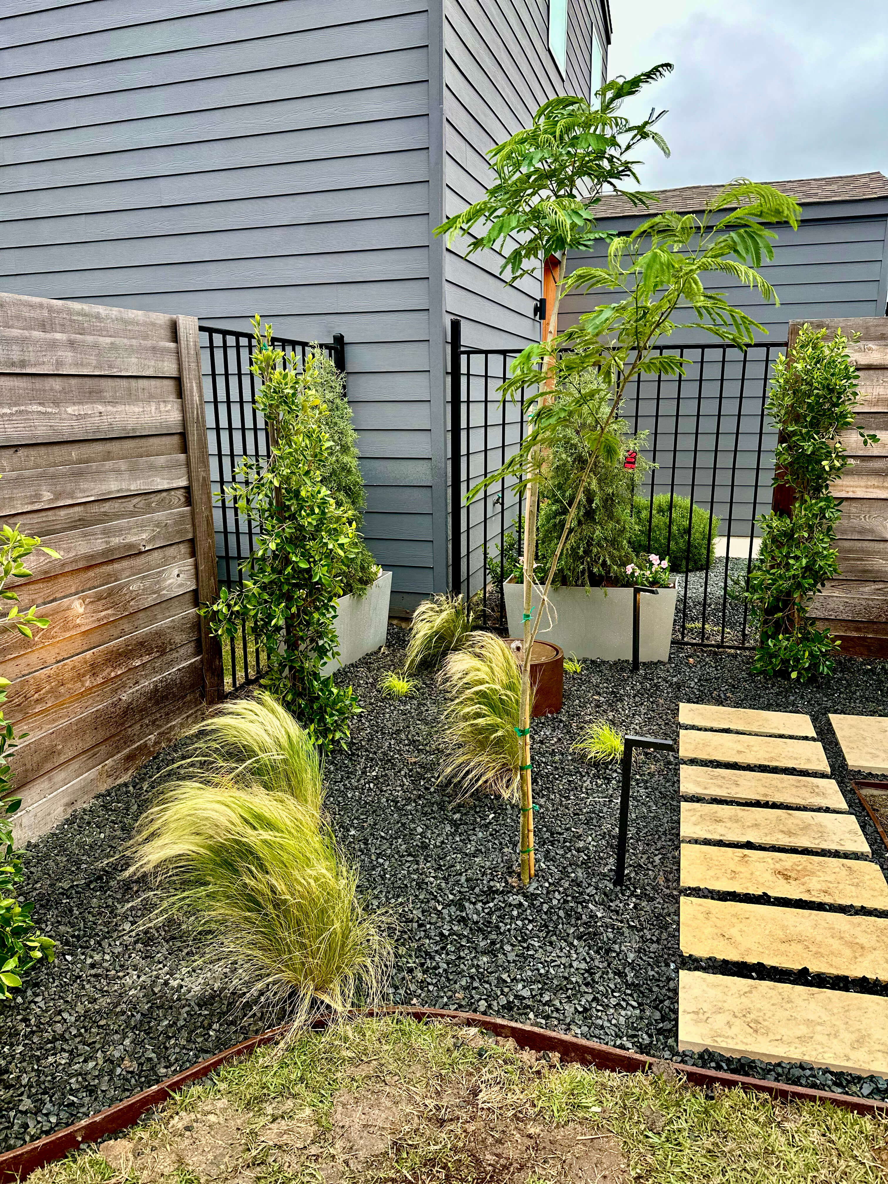 Modern small backyard landscape design with black gravel, privacy screening, pavers, planters, ornamental grasses, and Mediterranean-inspired planting