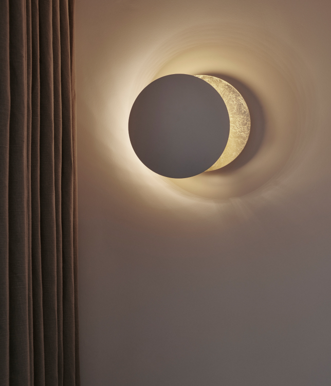 Wall-mounted light resembling a solar eclipse casting a warm glow on a plain wall next to brown curtains.