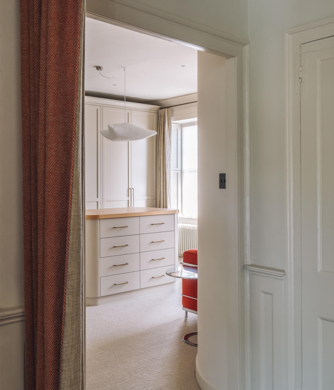 View through a doorway into a room with built-in cabinets, a hanging light fixture, and a red chair next to a glass side table.