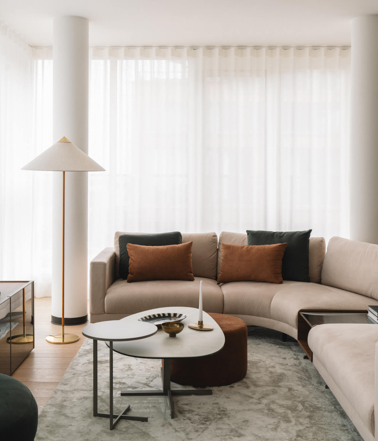 Modern living room with beige curved sofa, brown and dark green cushions, layered white coffee tables, floor lamp, and sheer white curtains.
