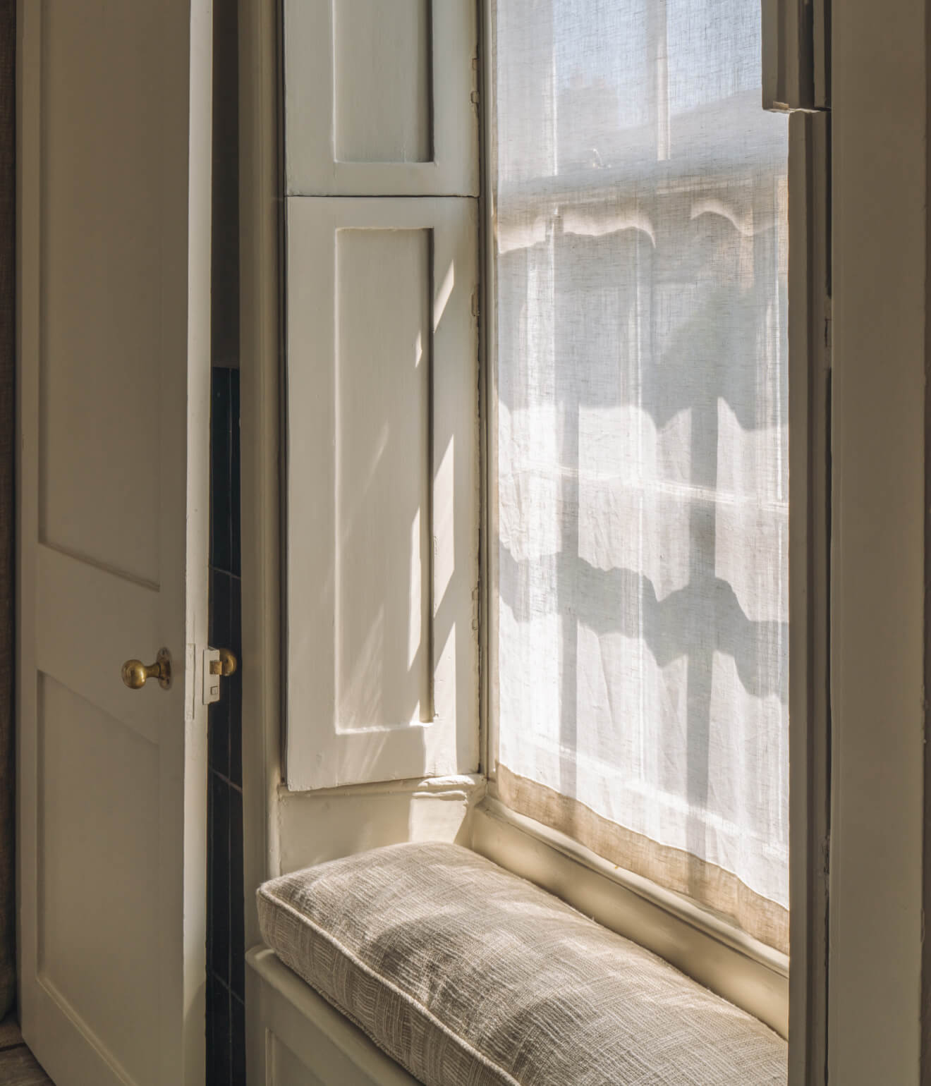 Sunlight casting shadows through a sheer curtain onto a cushioned window seat framed by white wooden panels and a partially open door with a brass knob.