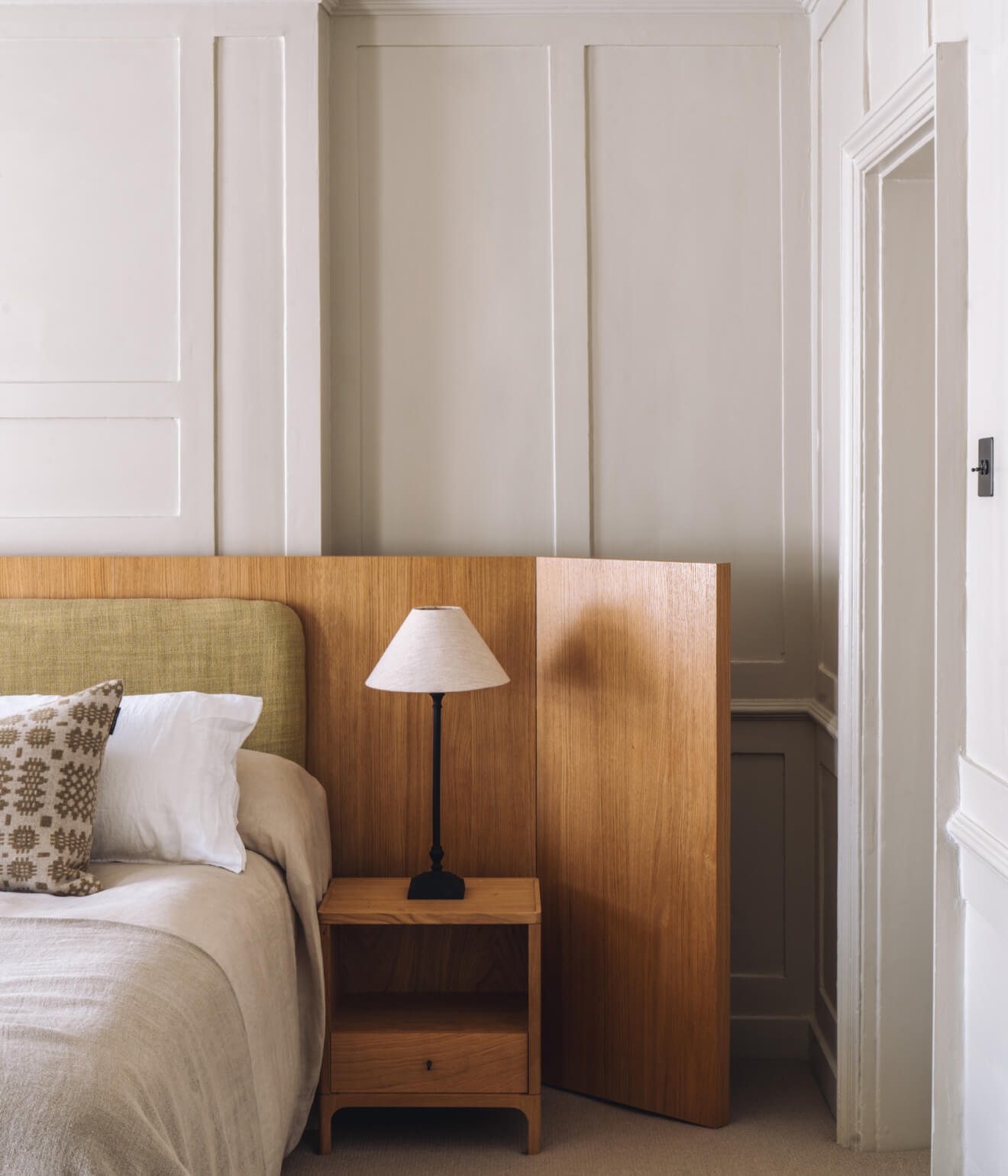 Cozy bedroom corner with beige bedding, patterned pillow, wooden nightstand, and lamp against paneled walls.
