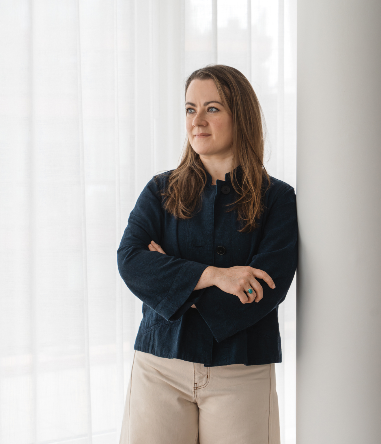 An image of Elizabeth Keenan, wearing a dark blue jacket and beige pants, standing with arms crossed near a white wall and sheer curtains.