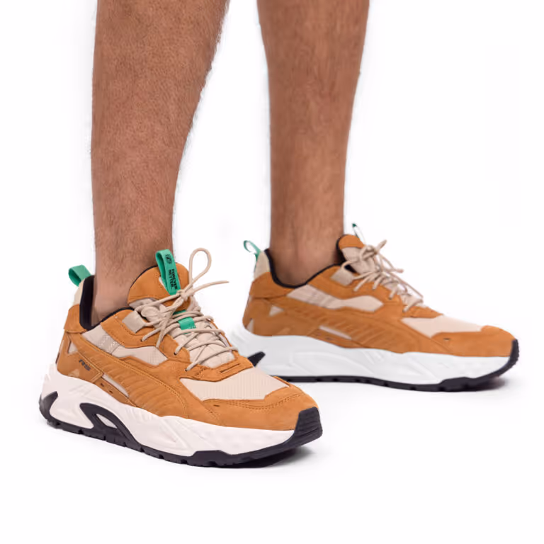 Pair of brown and beige athletic sneakers with green pull tabs worn on feet against white background.