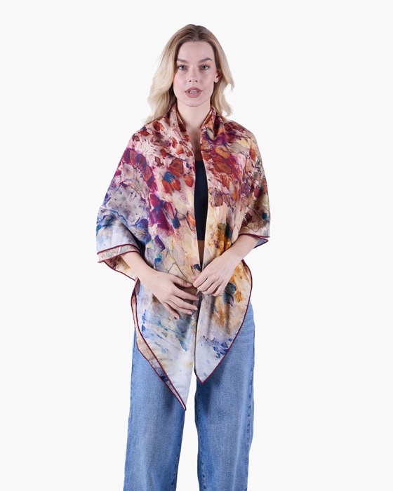 Woman wearing a multicolored scarf with an abstract pattern draped over her shoulders, paired with blue jeans and a black top on a white background.