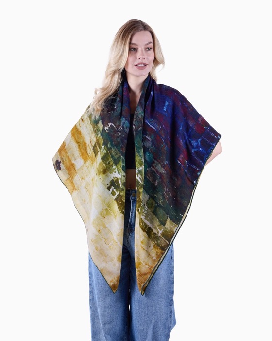 Blonde woman wearing a large rectangular scarf with an abstract diagonal pattern in shades of yellow, green, blue, and dark red, paired with high-waisted blue jeans and a black top, standing against a white background.