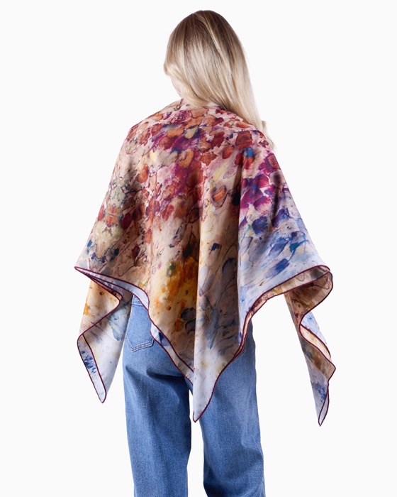 Person with long blonde hair facing away, wearing blue jeans and a colorful, watercolor-patterned scarf or wrap draped over their shoulders.