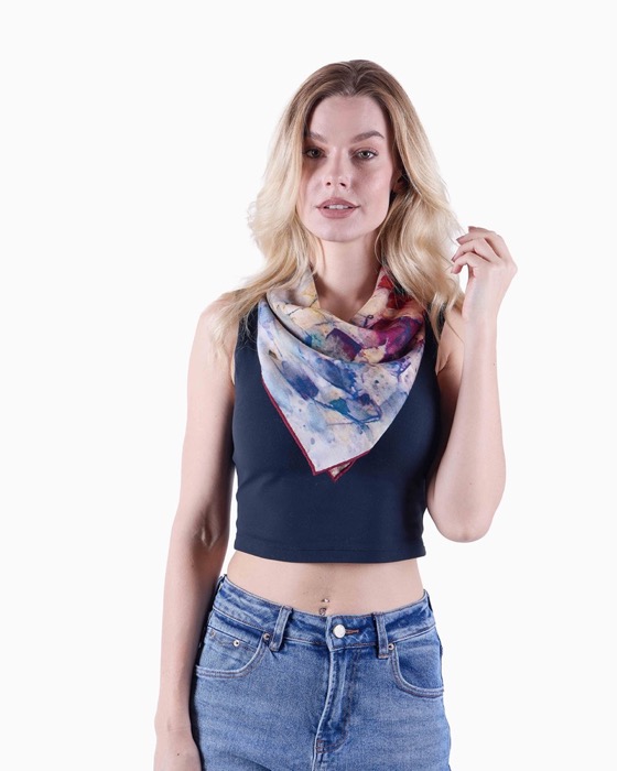 Blonde woman wearing a colorful patterned scarf tied around her neck, a black crop top, and blue jeans, standing against a white background.