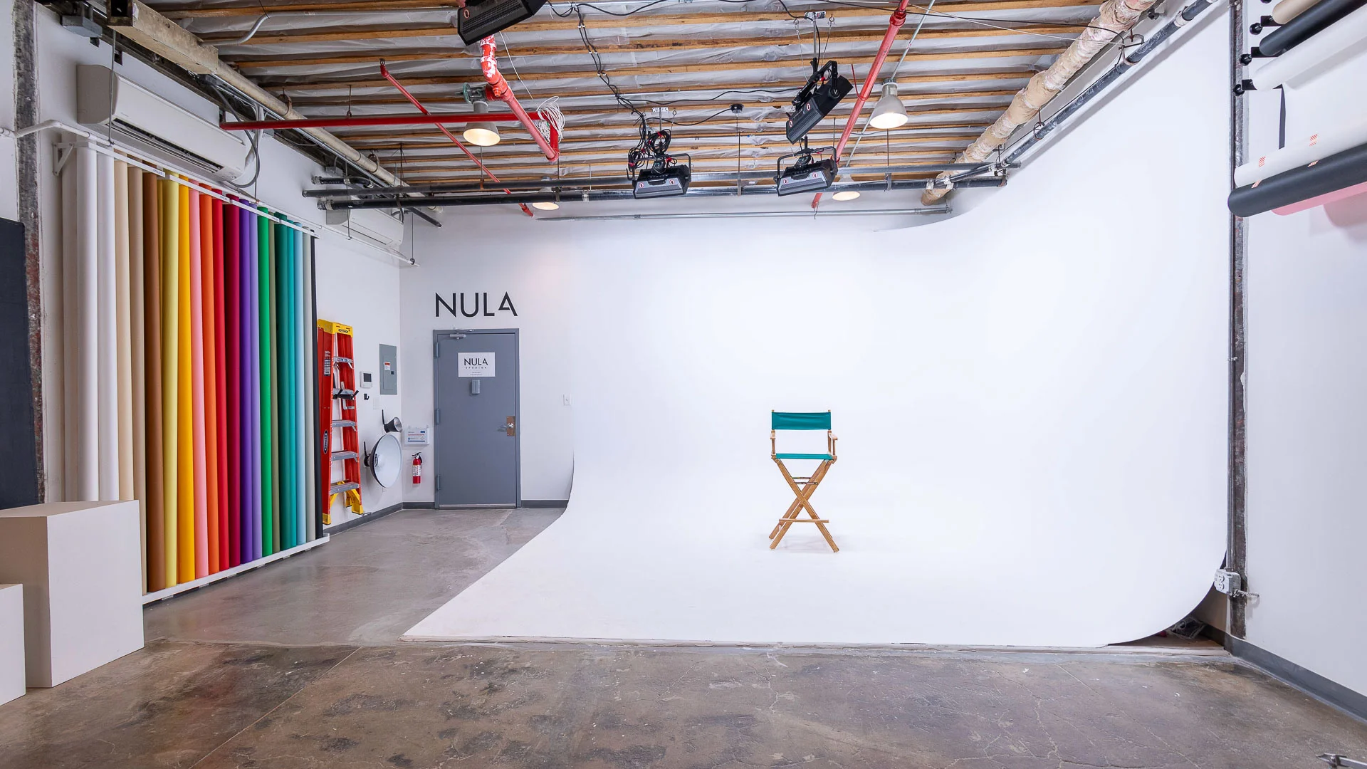 Bright photography studio with white cyclorama wall, a wooden director's chair with green fabric, colorful backdrop rolls on the left, ceiling lights, and exposed pipes.