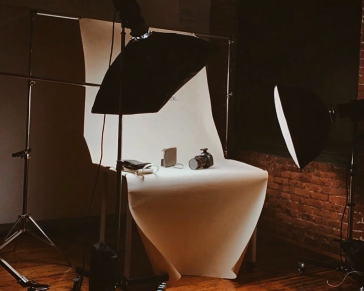 Small indoor photography studio setup with a table covered by a white backdrop sheet, surrounded by two large studio lights on stands, and a camera along with other small objects placed on the table.