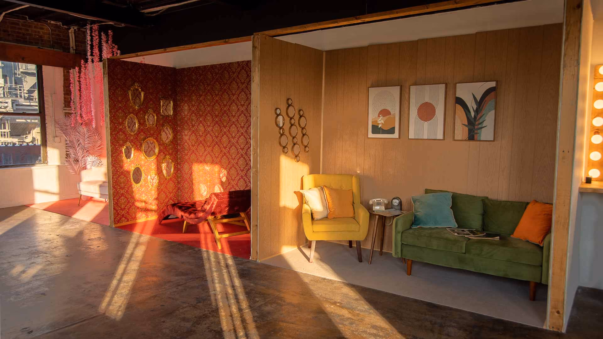 Sunlit loft interior with two cozy seating areas separated by partial wooden walls; one area features a yellow armchair with pillows and a green sofa with cushions and artwork above, the other has red wallpaper with gold patterns, a matching chair, and small mirrors on the wall.