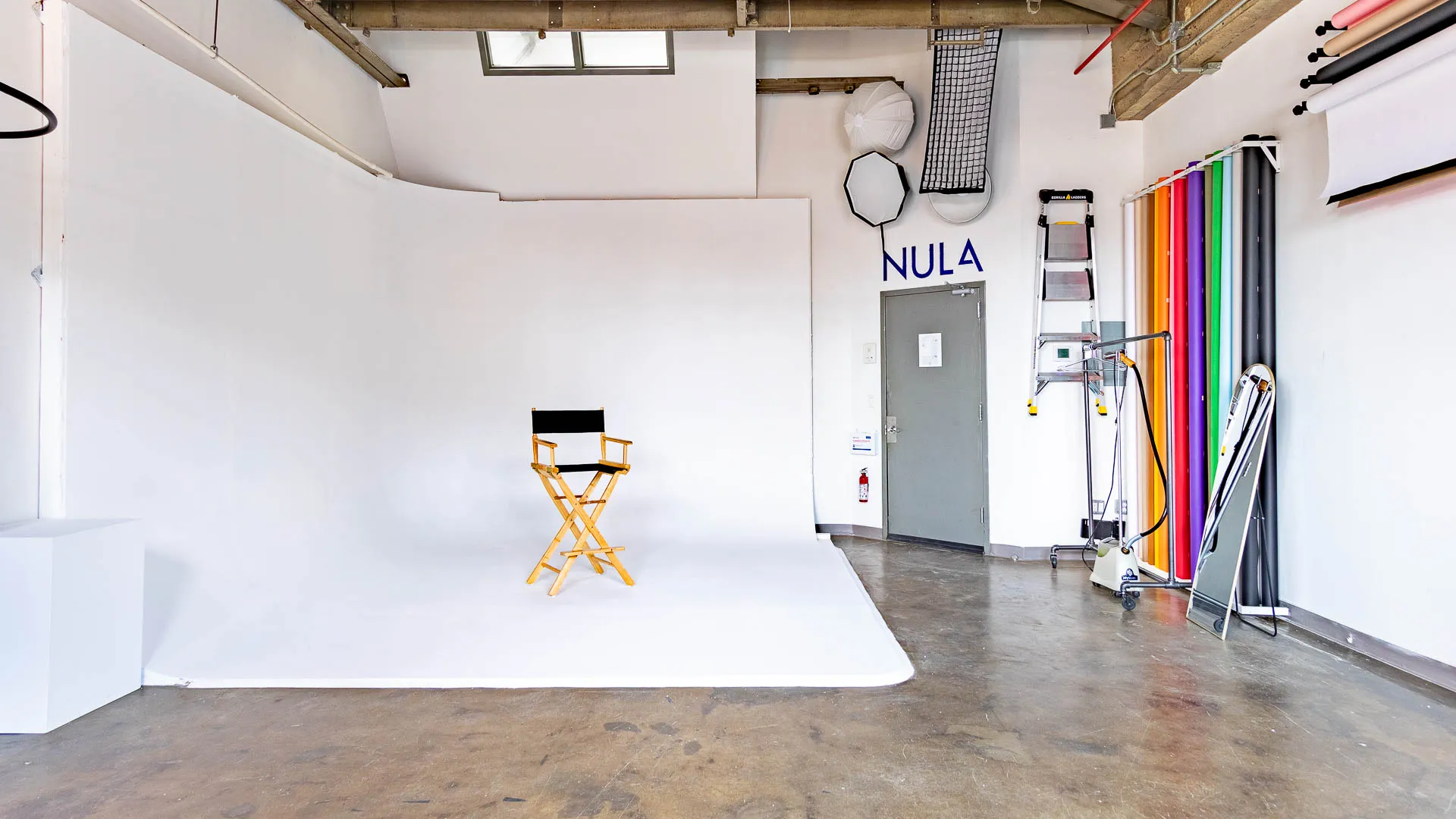 Bright photo studio with white cyc wall backdrop and a wooden director's chair with black seat in the center, colorful backdrop rolls, studio light modifiers, a ladder, and a tall mirror leaning against the wall.