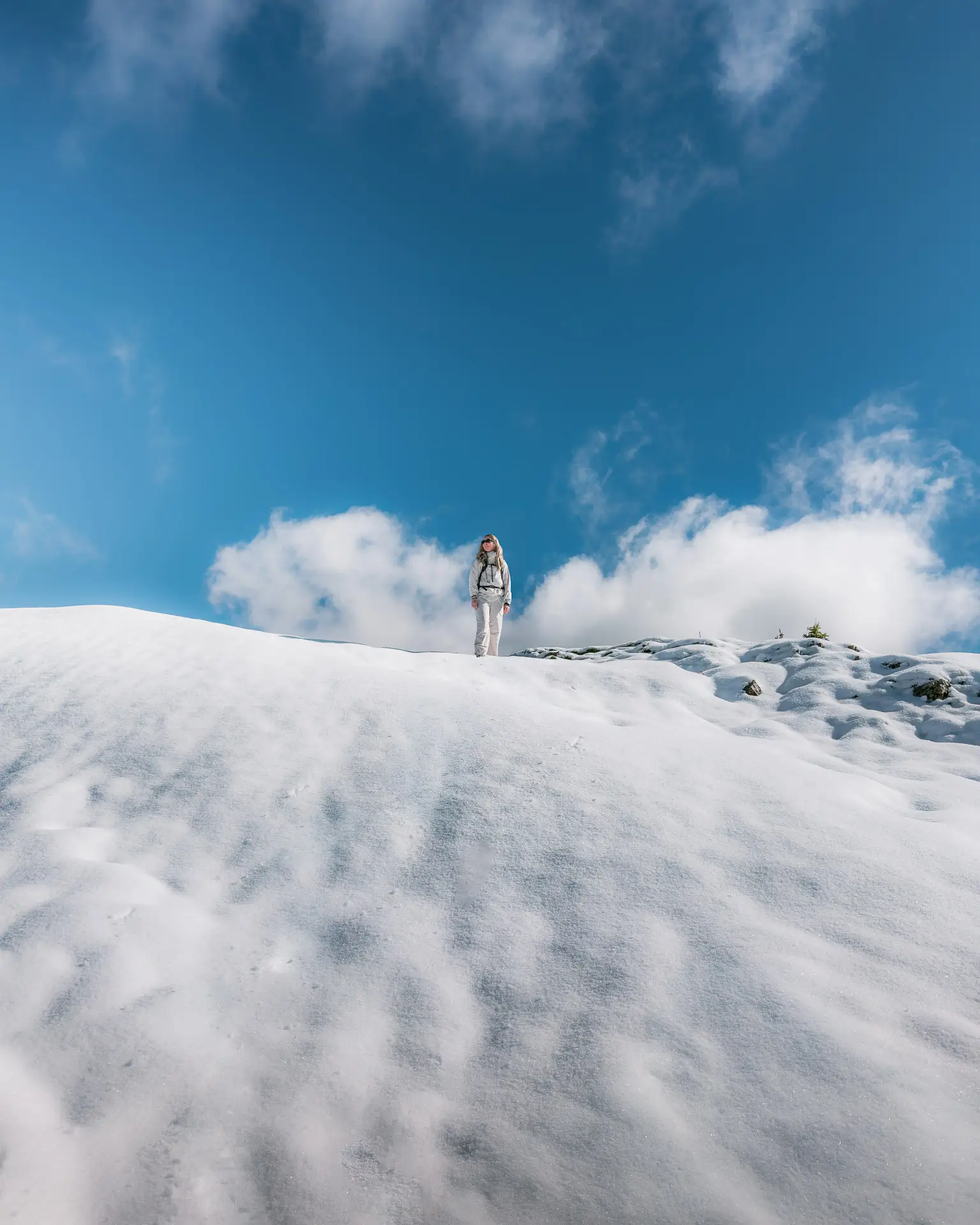 Hiker in white winter gear walking along a snow-covered ridge beneath a wide blue sky on Jumpingpound Summit Trail.