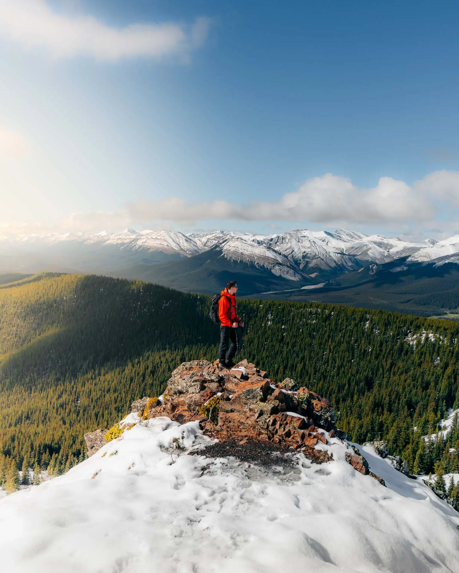 Hiker standing on a rocky outcrop at Jumpingpound Summit Trail overlooking snow-capped Kananaskis peaks at golden hour.