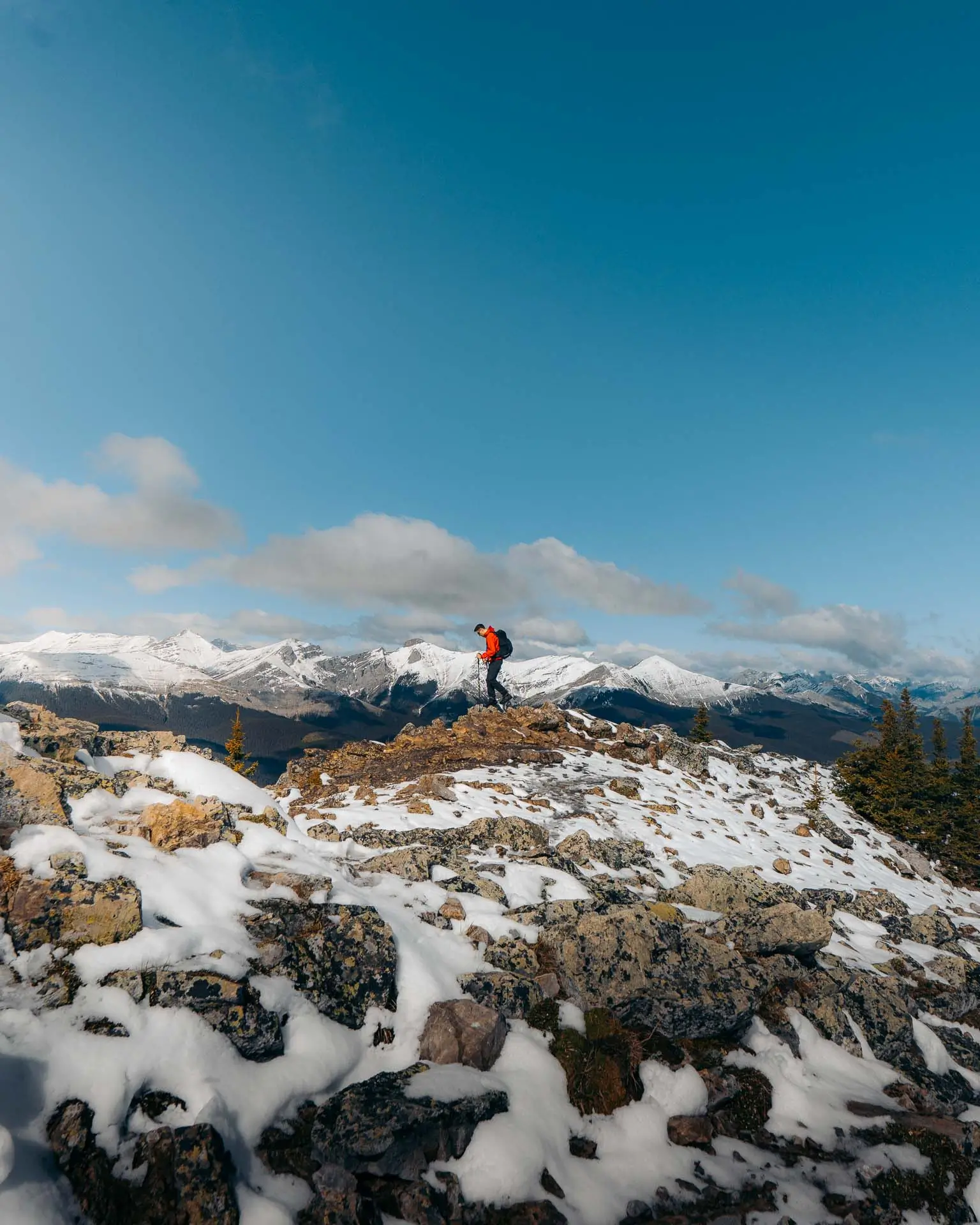 Hiker standing on a rocky ledge on Jumpingpound Summit Trail with snow-capped mountains stretching across the horizon.