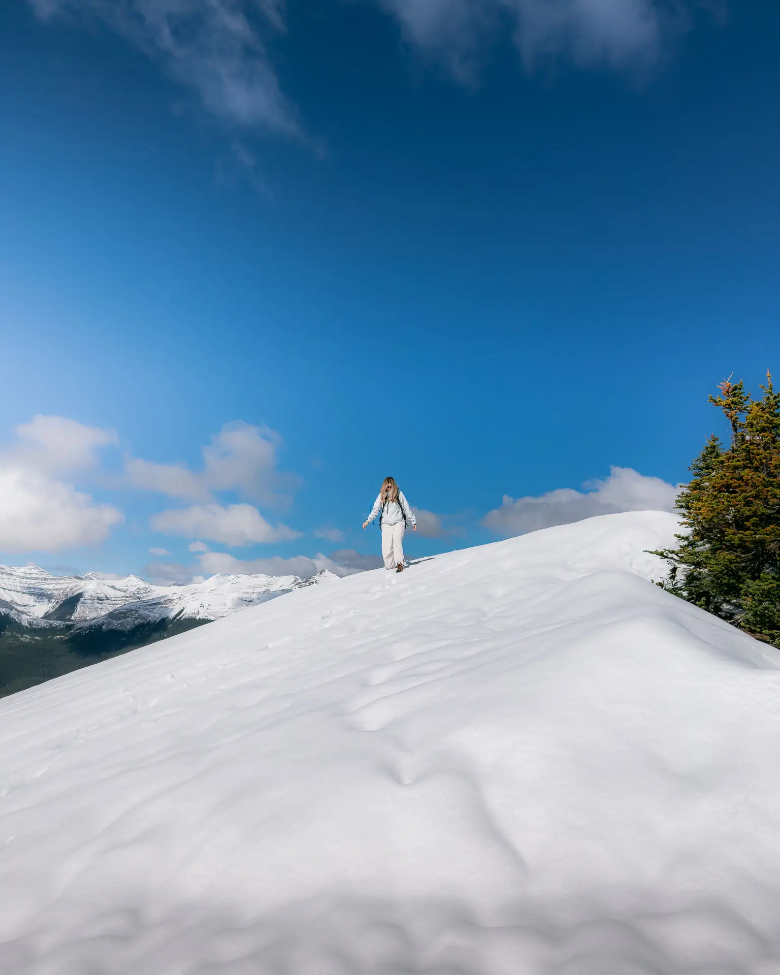 Hiker walking a snowy ridge on Jumpingpound Summit Trail with the Kananaskis mountain range visible in the distance.
