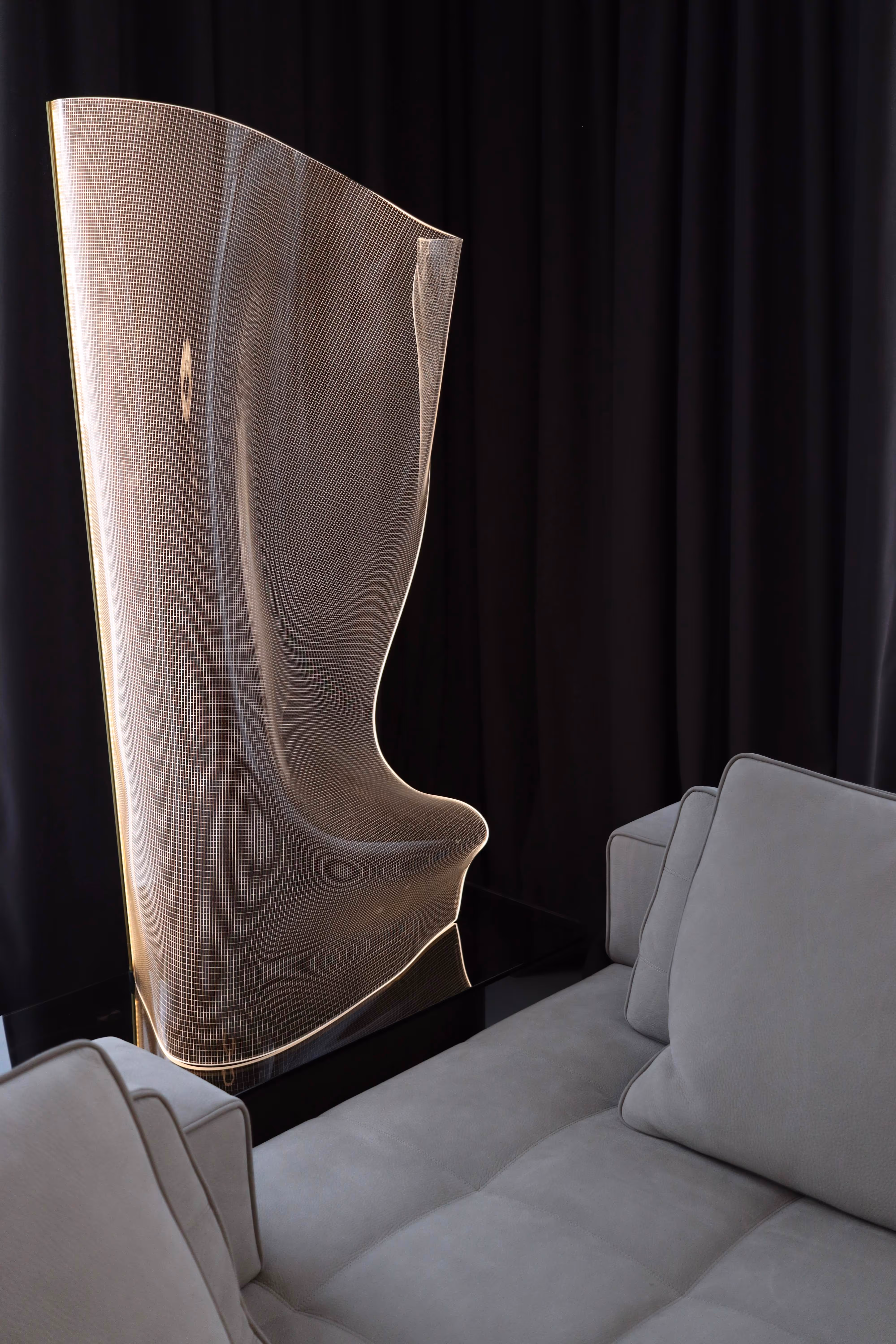 Modern translucent chair with curved, illuminated grid design next to a gray sofa with cushions.