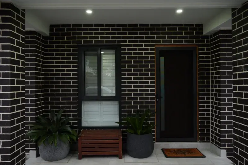 Covered front entry of the granny flat with dark brick walls and timber bench
