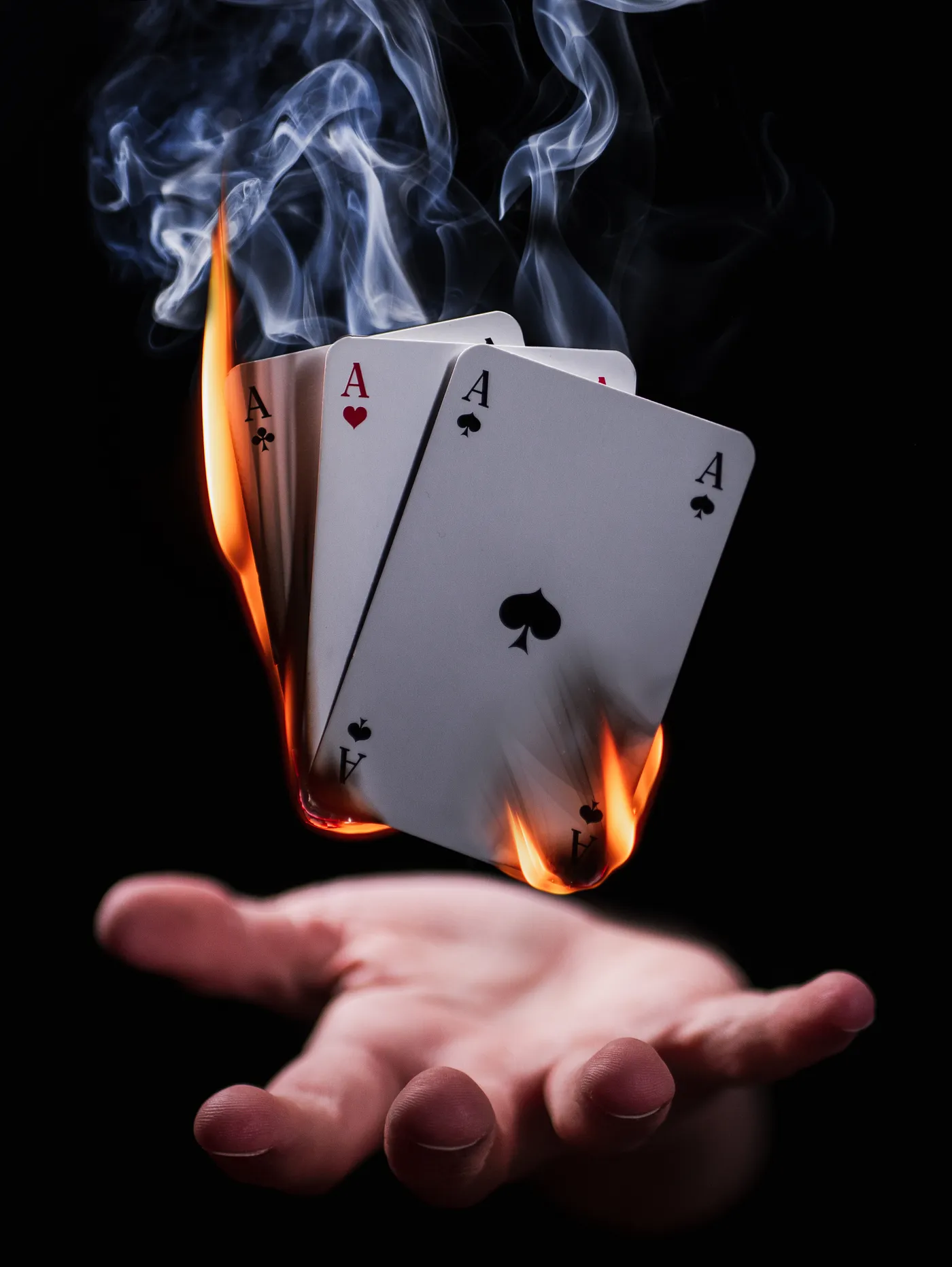 A dramatic image of a hand holding four aces, with the tops of the cards ablaze, smoke rising from the flaming cards against a dark background, symbolizing the overcoming of challenges and the ignition of action.