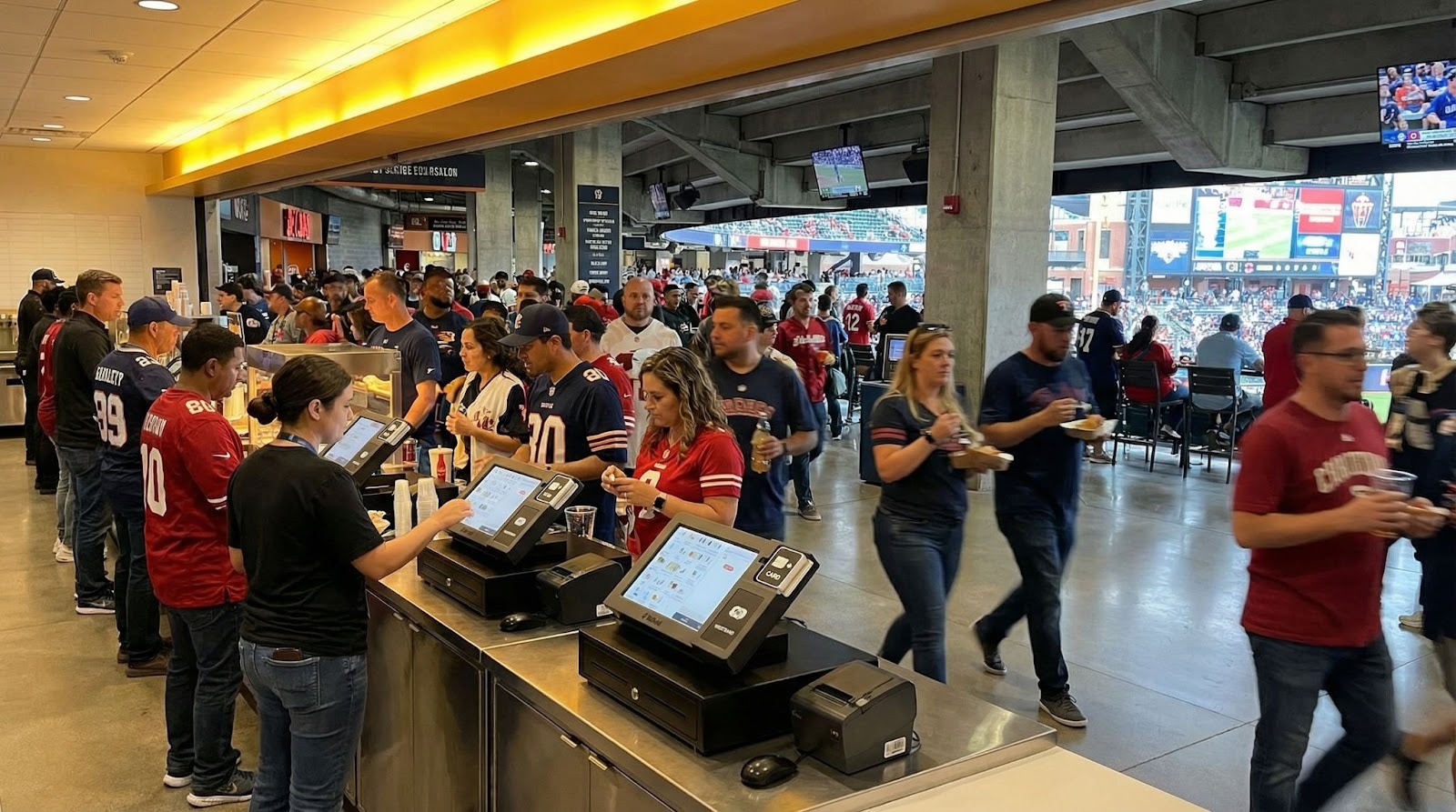 Busy stadium concourse with fans using touchscreen POS terminals at concession stands during a game day