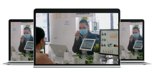 Two masked women sitting across a desk with computer screens displaying charts in an office with a social distancing sign.