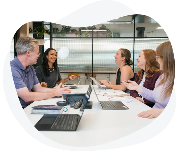Group of five people sitting around a table with laptops, smiling and engaged in a lively discussion in a modern office space.