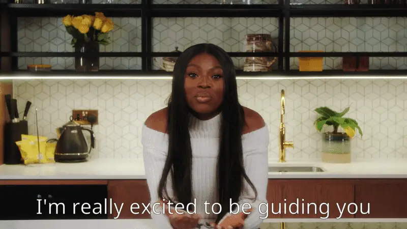 Woman with long dark hair wearing a white off-shoulder sweater speaking in a kitchen with a gold faucet and yellow flowers, text reads 'I'm really excited to be guiding you'.