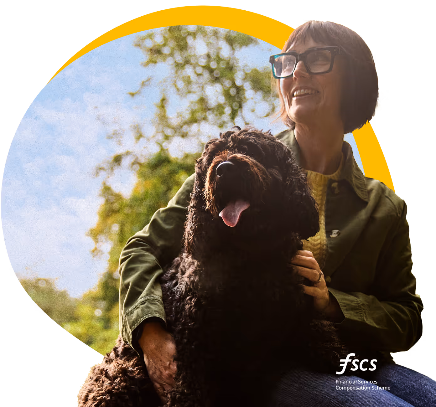  A woman with dark hair and glasses (featuring teal frames) is smiling brightly and looking up and to the right while sitting outdoors. She is wearing a green jacket and a yellow jumper. She is holding a large, dark brown, shaggy dog (possibly a Cockapoo or Labradoodle) on her lap, which is looking forward with its tongue hanging out. The background is a bright, slightly blurred view of a sunny park or forest, with green trees and a pale blue sky. A large, yellow, abstract oval shape frames the image content behind the woman's head and shoulders. The logo "fscs Financial Services Compensation Scheme" is visible in the bottom right corner.