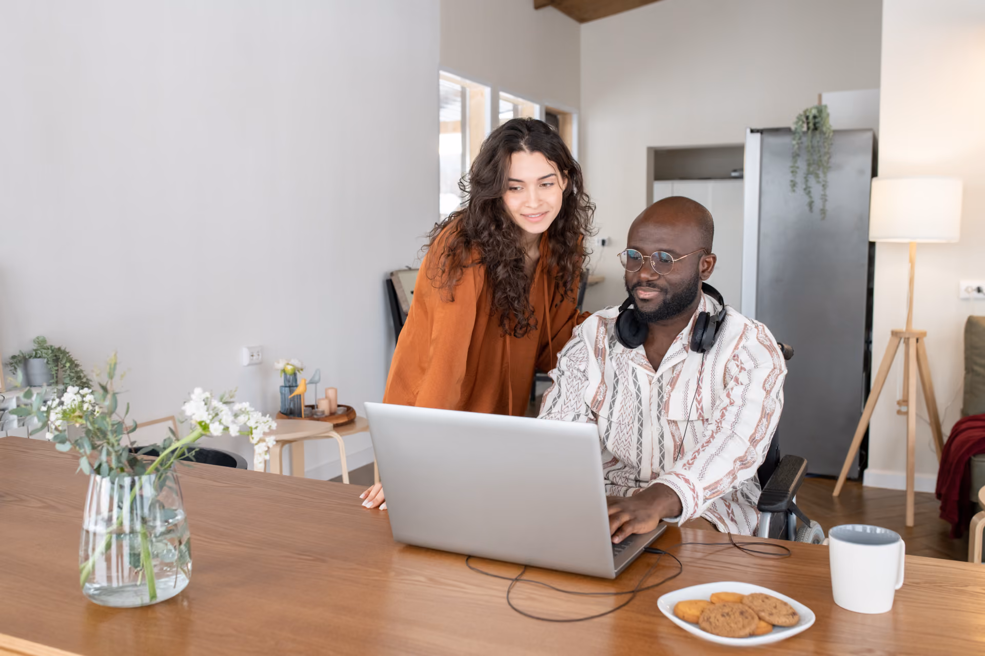 Man in wheelchair and woman looking at a laptop together at a wooden table with cookies and a mug.