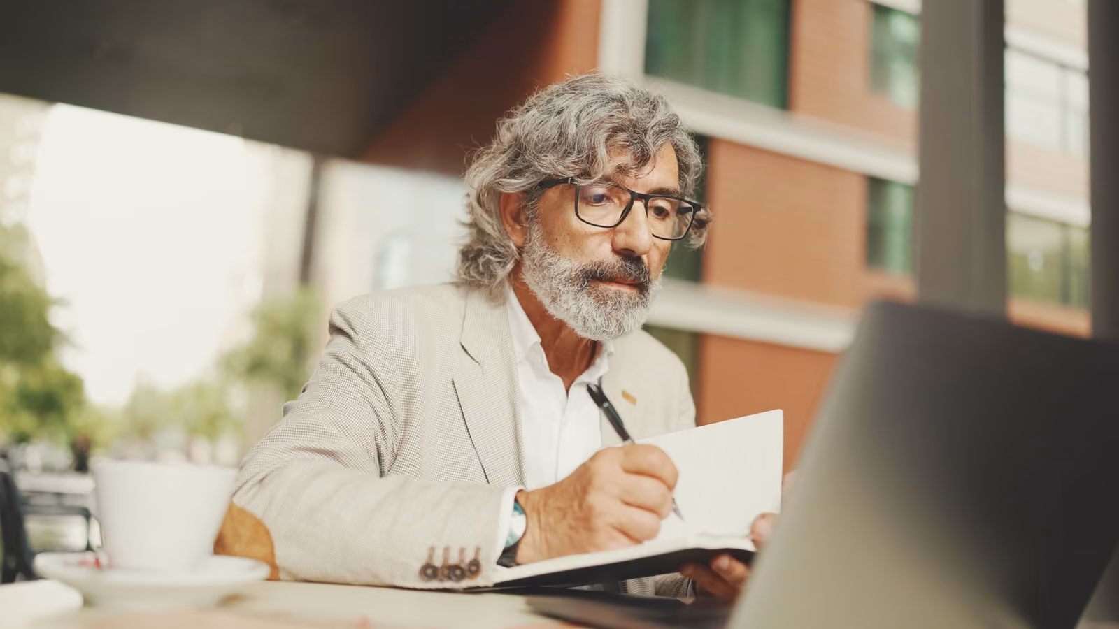 Older man with gray hair and beard writing in notebook while working on laptop outdoors.