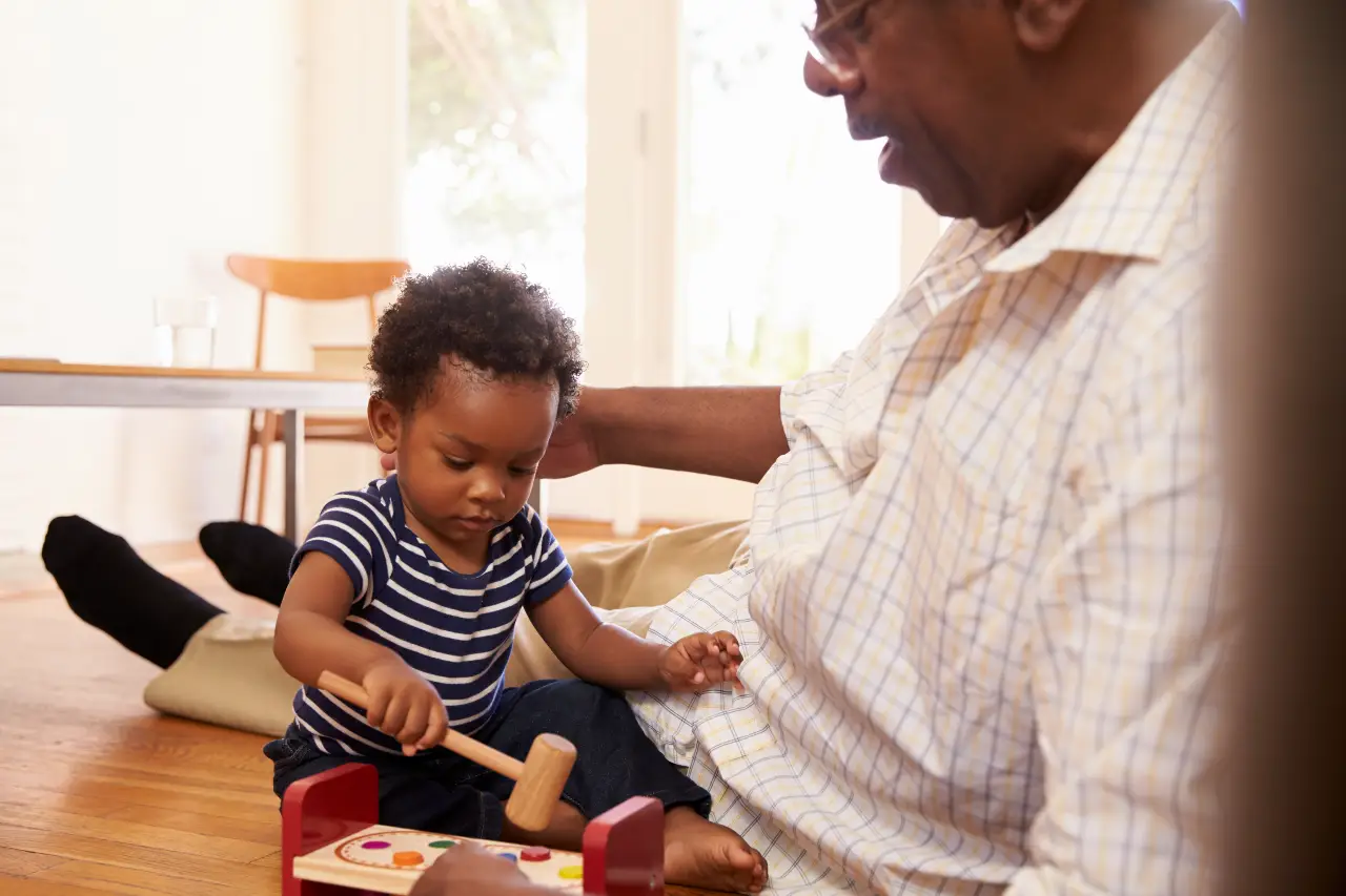 Grandfather sitting on floor playing with toddler who is using a wooden toy hammer.