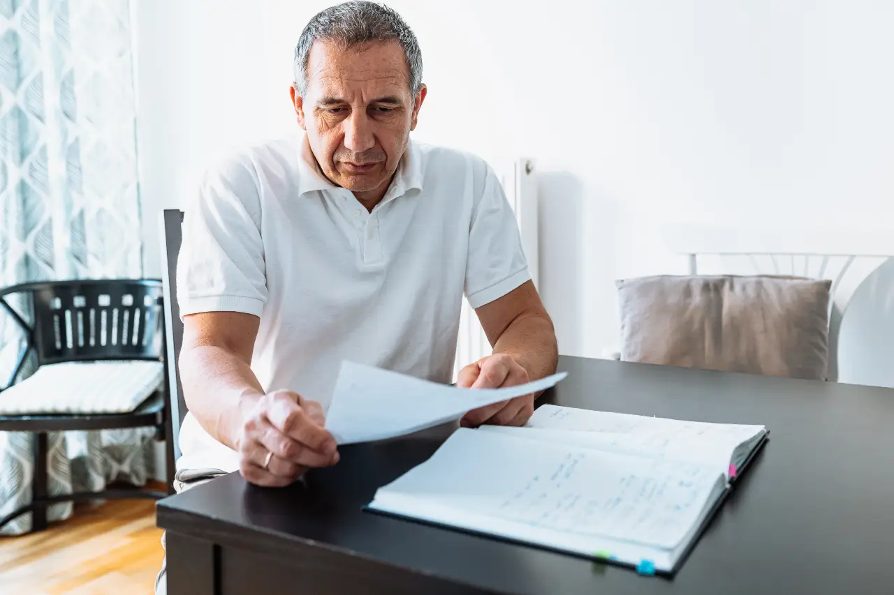 Middle-aged man in white polo shirt reads documents at a black table with a notebook.