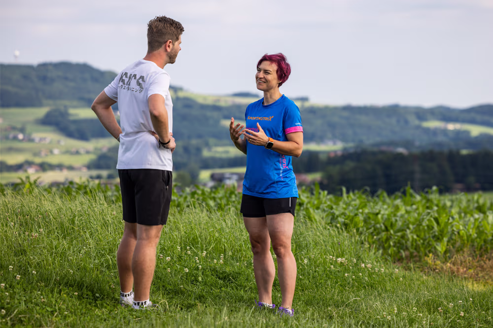 Zwei Sportler in Sportkleidung unterhalten sich auf einer Wiese mit hügeliger Landschaft im Hintergrund.