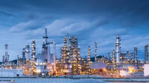 Large industrial oil refinery complex with tall distillation towers illuminated against a cloudy evening sky.