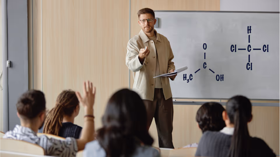 Male chemistry teacher explaining molecular formulas on whiteboard to students in a classroom.