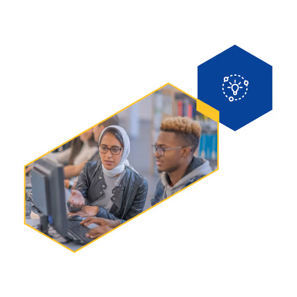 Two students, one wearing a hijab and glasses, collaborate and discuss while using a desktop computer in a library setting.