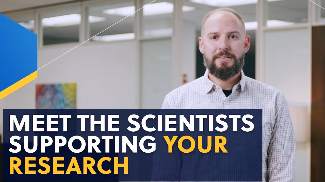 Bearded man in a checkered shirt standing in an office with the text 'Meet the scientists supporting your research' in white and yellow letters.