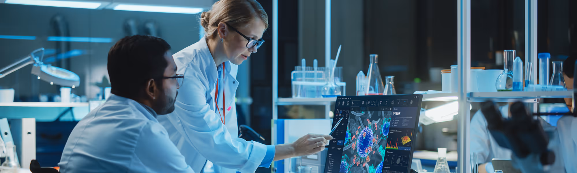 Two scientists in white lab coats examining a 3D virus model on a computer screen in a research lab