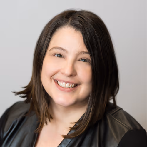 Portrait of a smiling woman with shoulder-length brown hair wearing a gray jacket, against a plain gray background.
