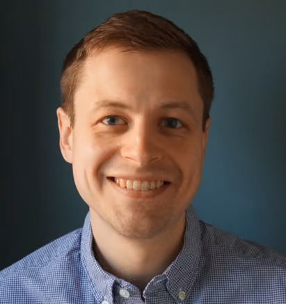 Smiling man with short brown hair wearing a blue checkered button-up shirt against a dark teal background.