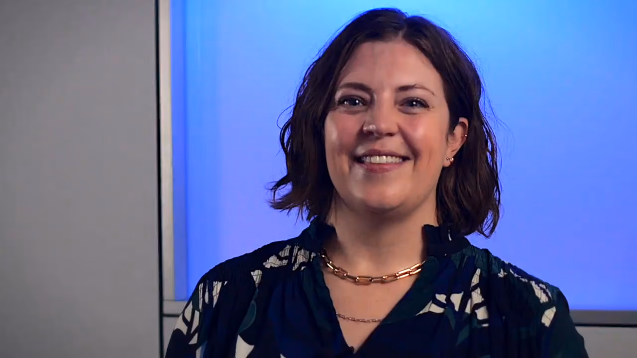 Smiling woman with short brown hair and gold chain necklace against a blue background.
