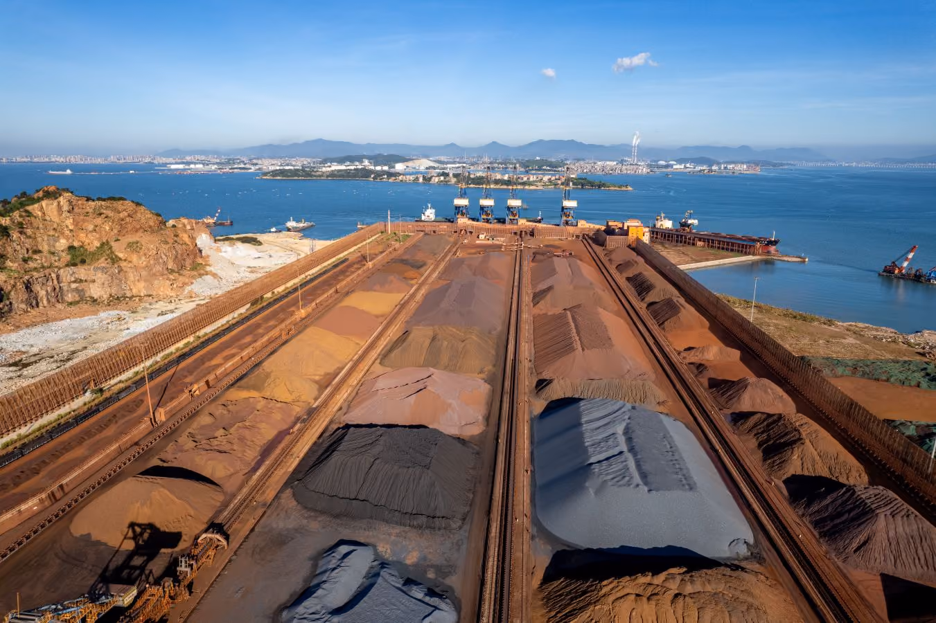 Aerial view of a large industrial port with multiple mounds of bulk minerals stored in long rows near water.