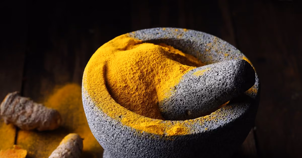 Gray stone mortar and pestle with bright yellow turmeric powder on dark wooden surface.