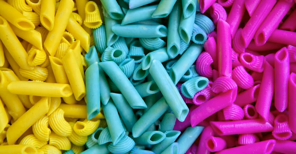 Yellow, teal, and magenta colored uncooked pasta in different shapes packed closely together.
