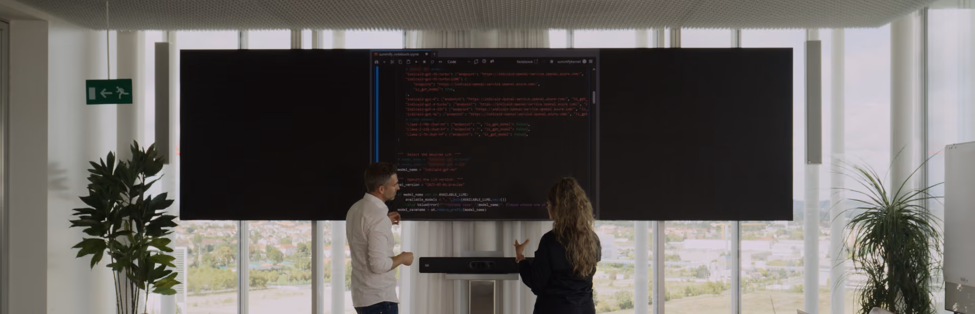 Two people reviewing code displayed on a widescreen monitor in a modern office with large windows.