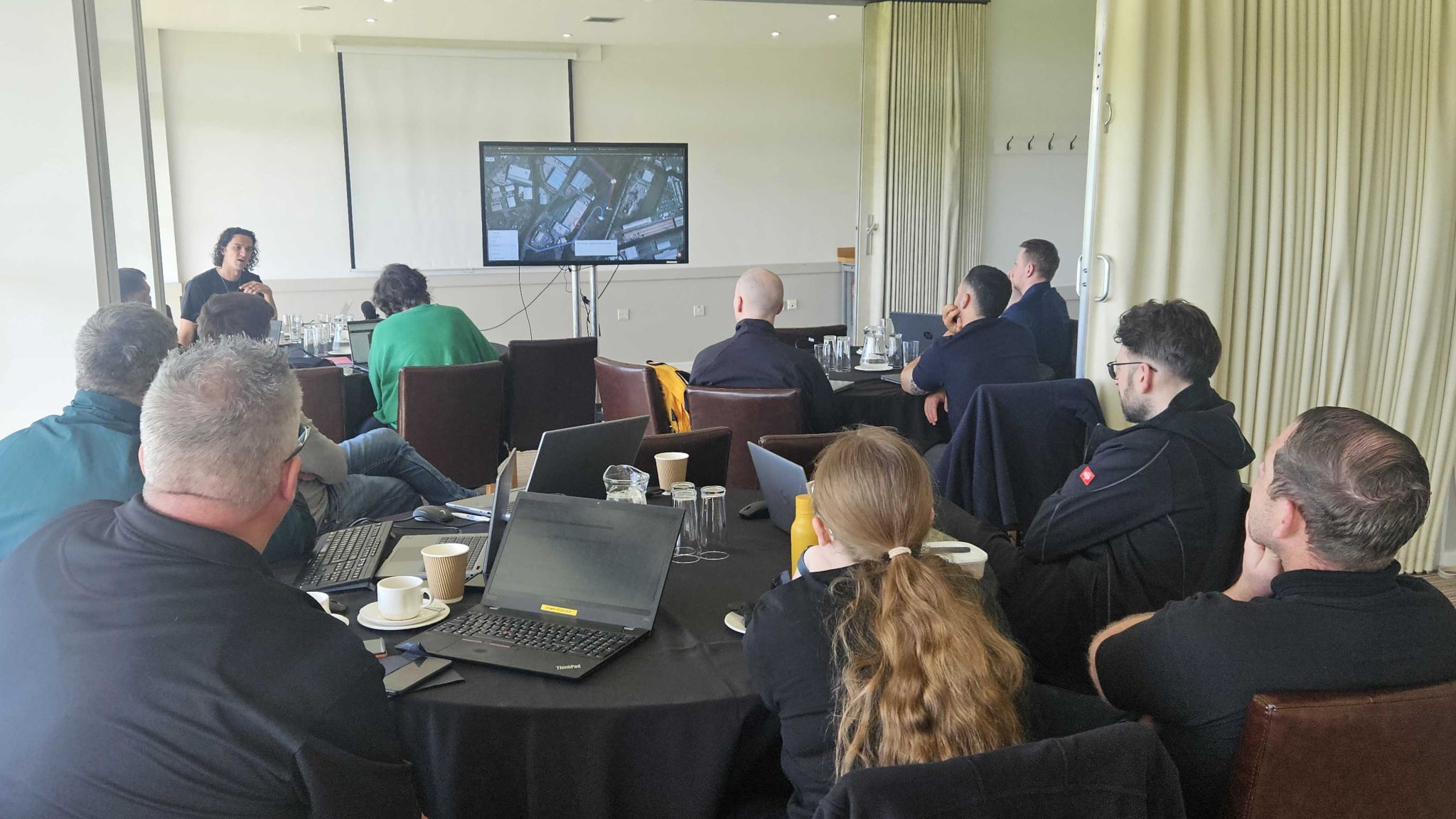 Project team at round tables with laptops, taking part in a hands-on software training session led by a presenter at the front.