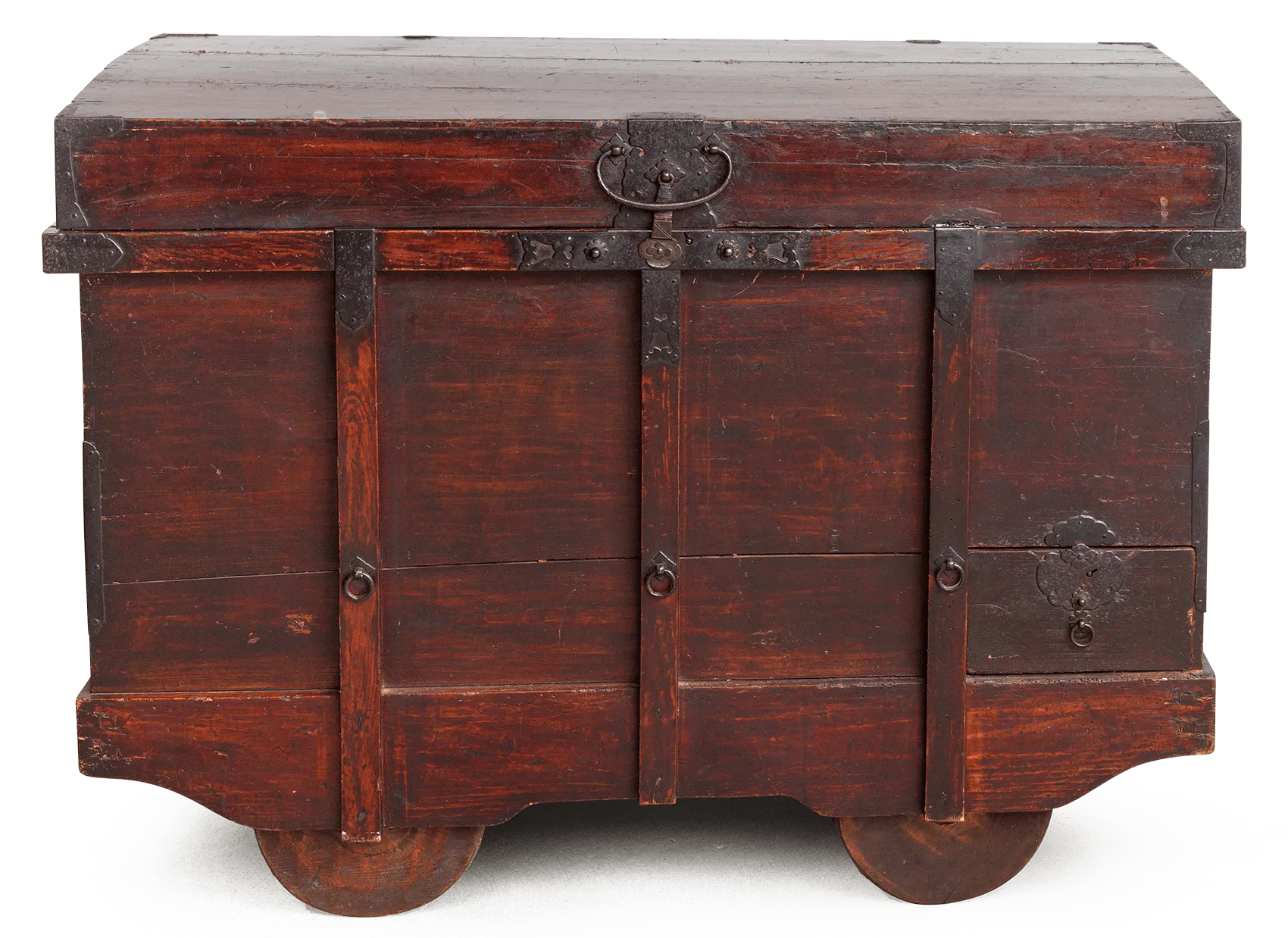 Große, dunkelrot gebeizte Holztruhe mit kräftigen Eisenbeschlägen und breiten vertikalen Verstärkungsbändern. Oben mittig ein massives Schloss mit Ringgriff, seitlich zusätzliche Ringgriffe; rechts eine kleine integrierte Schublade mit Zierbeschlag. Der Korpus steht auf zwei großen, scheibenförmigen Holzrädern