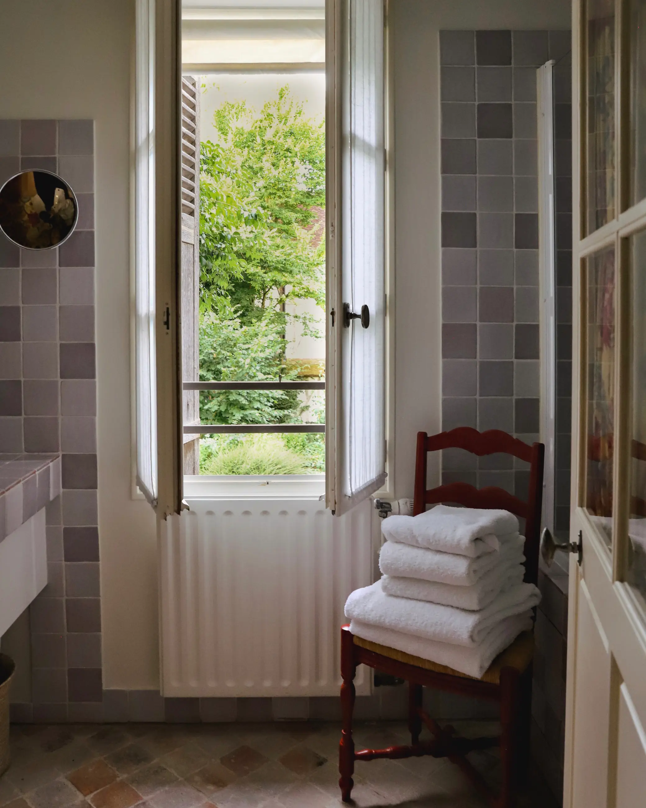 Open bathroom window in a suite at La Maison de Rhodes in Troyes, France, with soft natural light, lavender-toned tiles, and neatly folded white towels on a wooden chair