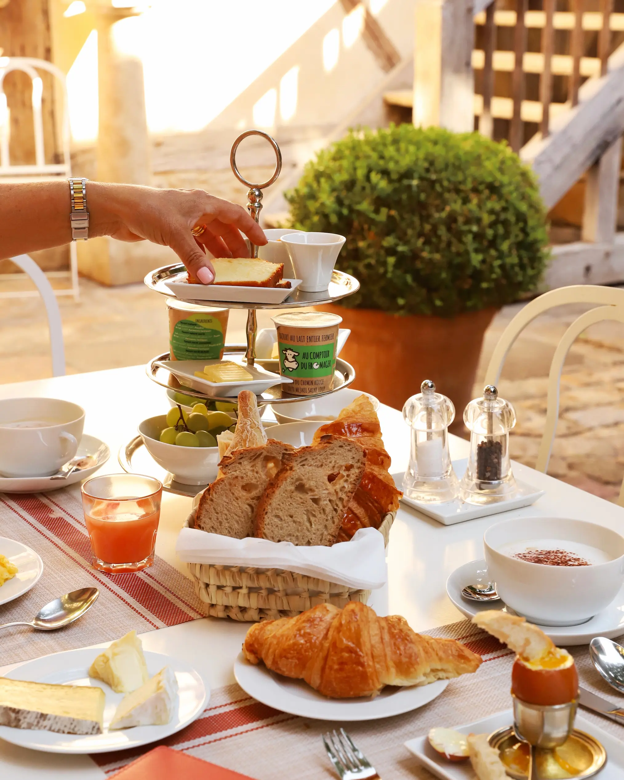 Elegant French breakfast served in a sunlit courtyard at La Maison de Rhodes, with croissants, fresh bread, cheese, yogurt, soft-boiled egg, juice, and coffee on a white table with striped linen