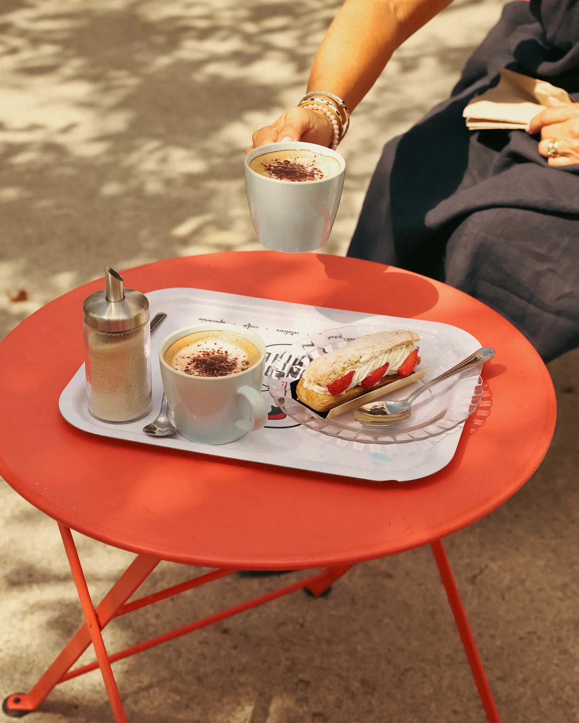 Outdoor café scene at Tonton Marine bakery in Troyes, France, with cappuccinos and a fresh strawberry éclair served on a red bistro table in dappled sunlight